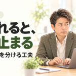 同僚に見られながらパソコンを操作し、手を止める男性。見られると、手が止まる。操作と説明を分ける工夫。